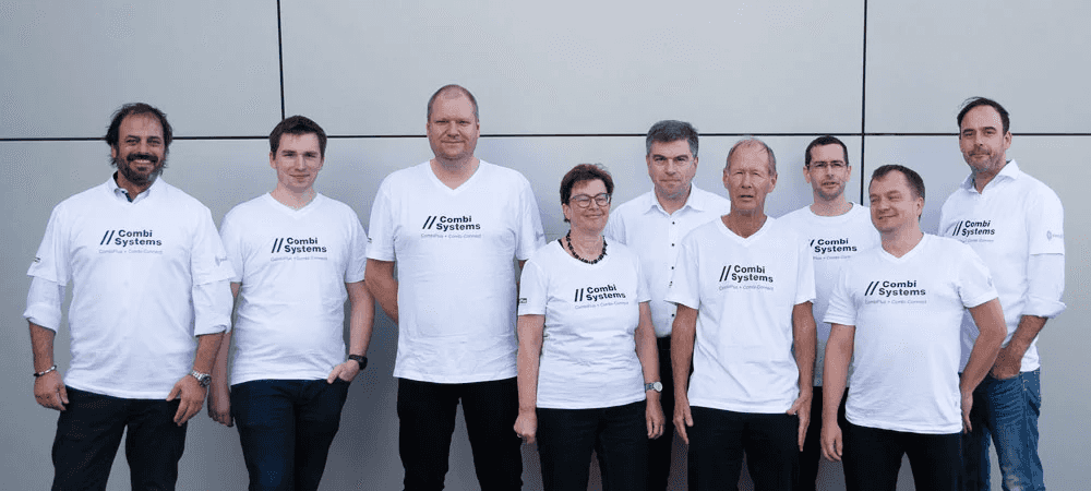 Gruppenfoto des CombiTeams vor einer grauen Wand, bestehend aus neun Personen in weißen CombiSystems-Shirts, die gemeinsam in die Kamera lächeln.