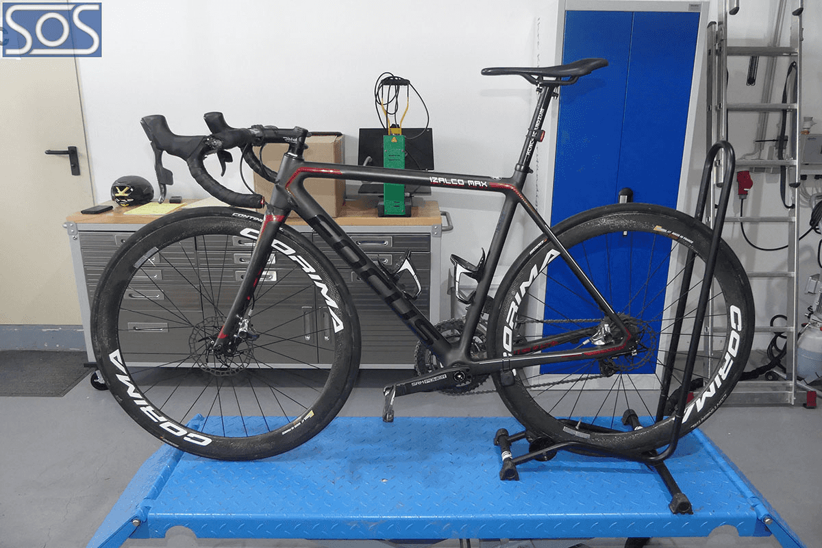 Ein schwarzes Rennrad der Marke Focus mit Carbonrahmen und Corima-Laufrädern steht zur technischen Überprüfung auf einem blauen Montageständer in einer Werkstattumgebung.