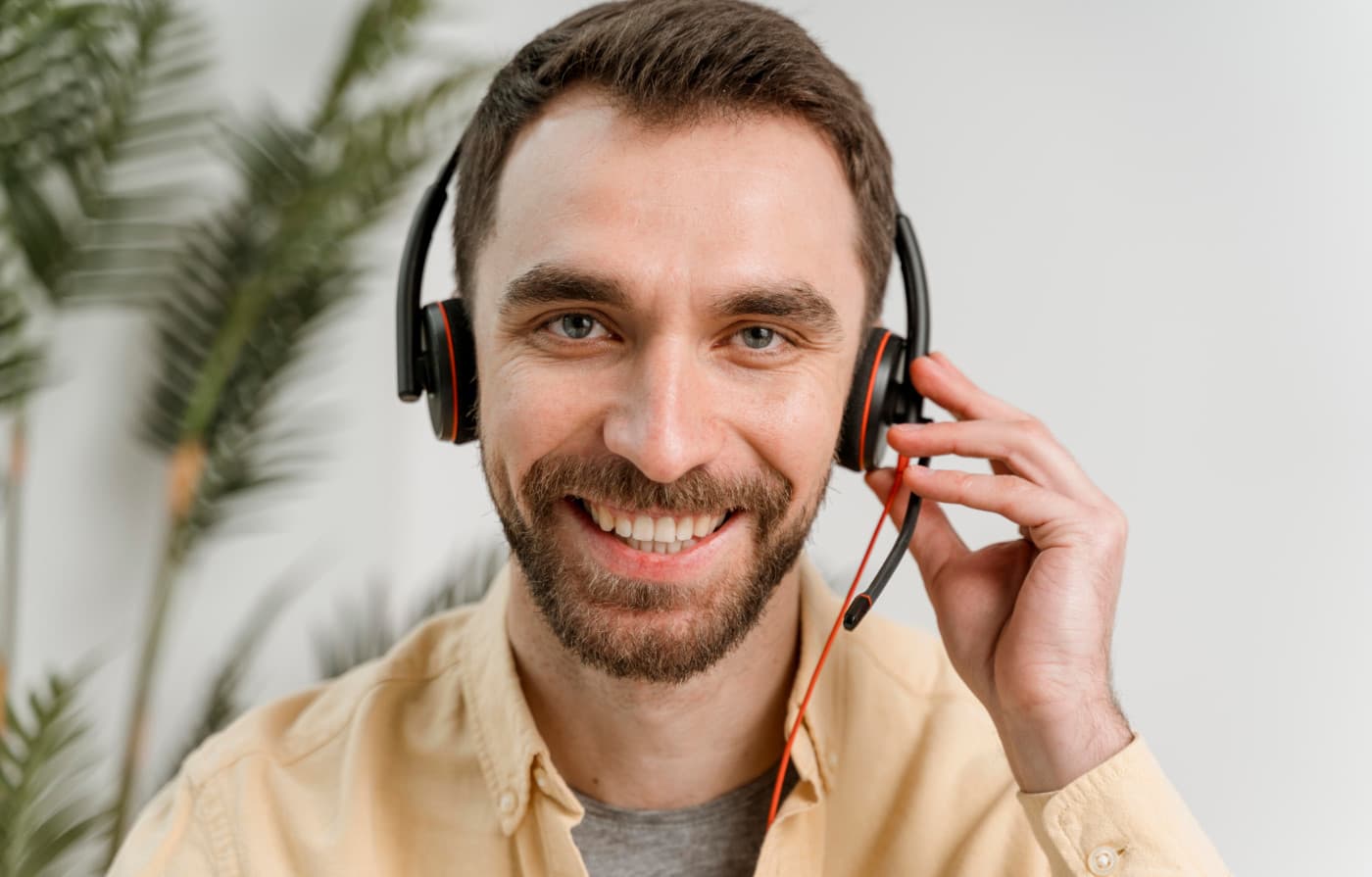 Einen Mann, der ein Headset trägt und in die Kamera lächelt, als würde er an einem Videoanruf teilnehmen. Er scheint sich in einer professionellen, aber entspannten Umgebung zu befinden. Der Hintergrund zeigt Pflanzen, was auf eine freundliche und angenehme Atmosphäre hinweist. Der Mann trägt ein beiges Hemd und sieht aus, als würde er in einem virtuellen Meeting oder einer Besprechung teilnehmen. Die Szene vermittelt eine moderne Arbeitsumgebung, die auf digitale Kommunikation und Zusammenarbeit setzt.