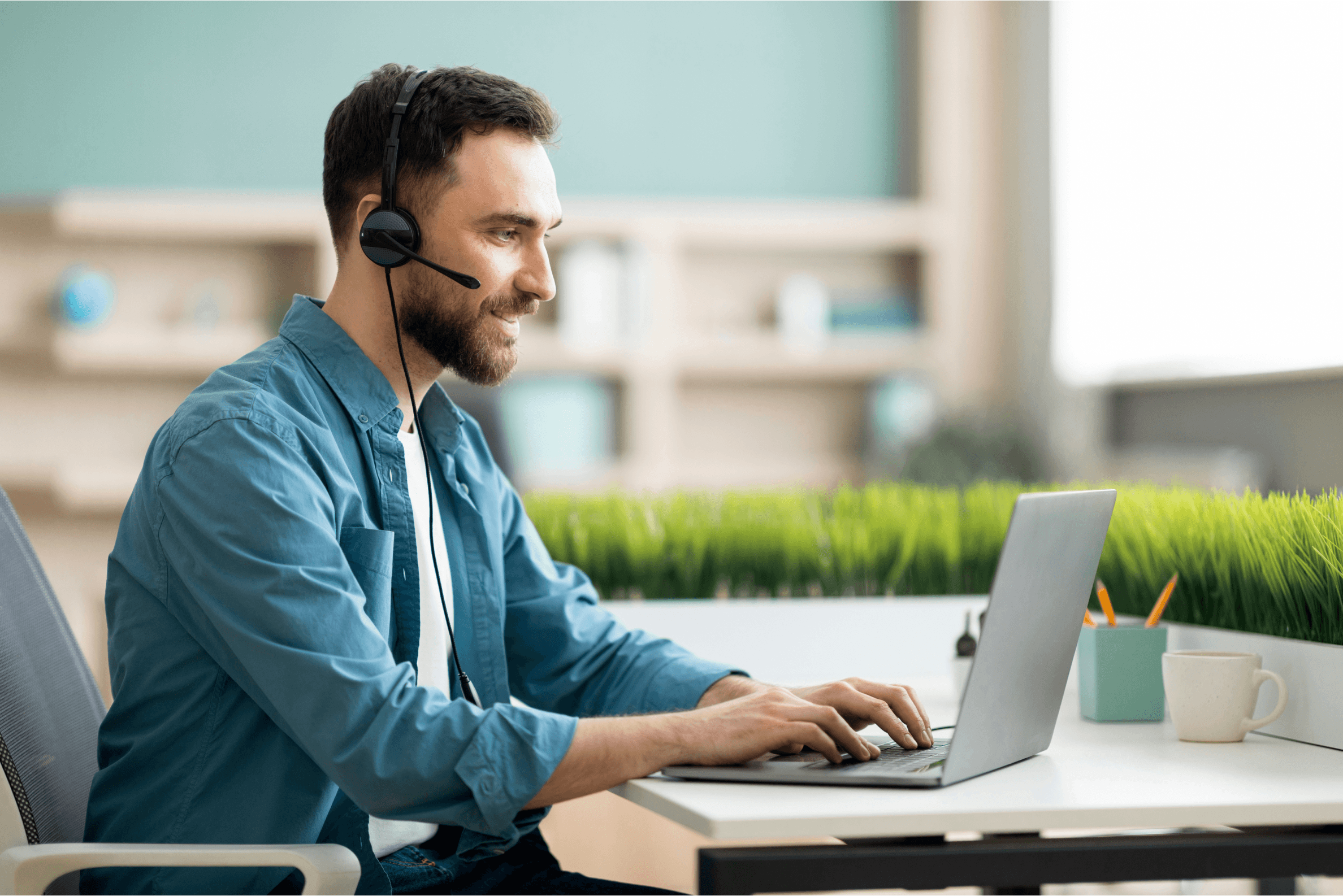 Das Bild zeigt einen Mann, der in einem modernen Büro mit einem Laptop arbeitet und ein Headset trägt. Er ist in der Mitte des Bildes abgebildet und konzentriert sich auf den Bildschirm, während er mit den Händen auf der Tastatur tippt. Der Hintergrund zeigt einen gut organisierten Raum mit Regalen, die Bücher und Dekorationsobjekte enthalten. Eine grüne Pflanze steht auf dem Tisch und verleiht dem Bild eine natürliche Atmosphäre.