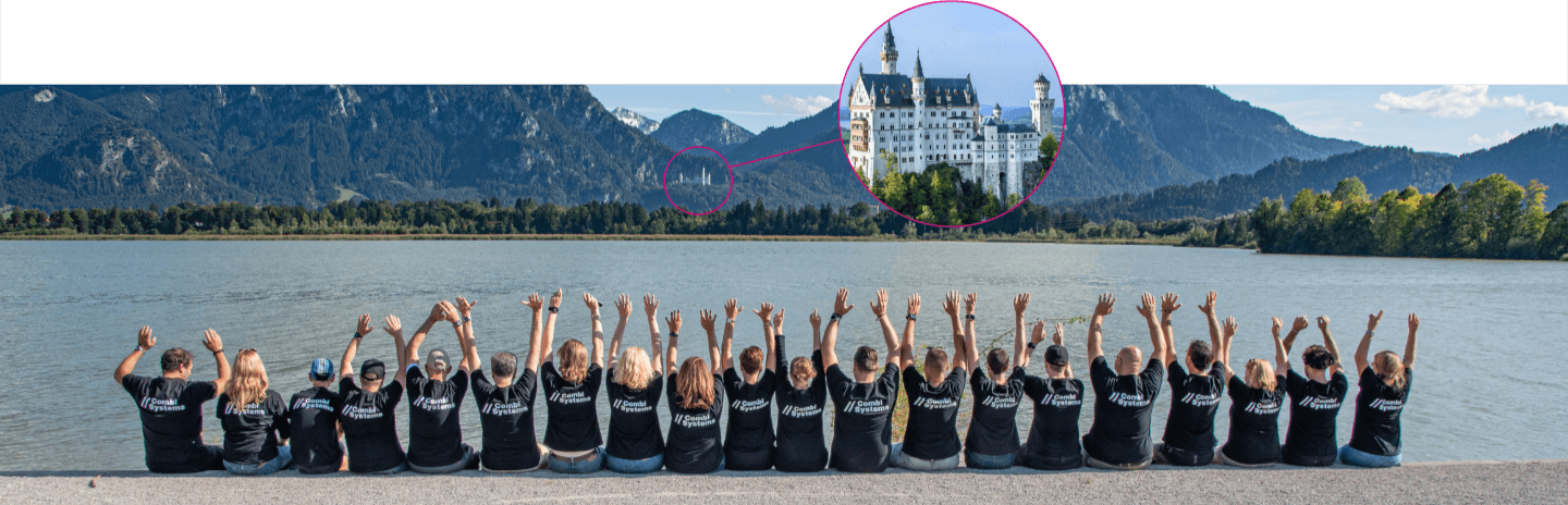 Das CombiTeam sitzt in einer Reihe mit dem Rücken zur Kamera vor einem malerischen See, umgeben von Bergen. Im Hintergrund ist Schloss Neuschwanstein zu sehen. Alle Teammitglieder tragen schwarze T-Shirts mit dem Logo von CombiSystems und heben gemeinsam die Hände in die Luft. Als Team arbeiten wir jeden Tag daran, unseren CombiKunden die Arbeit als Kfz-Sachverständiger zu erleichtern.