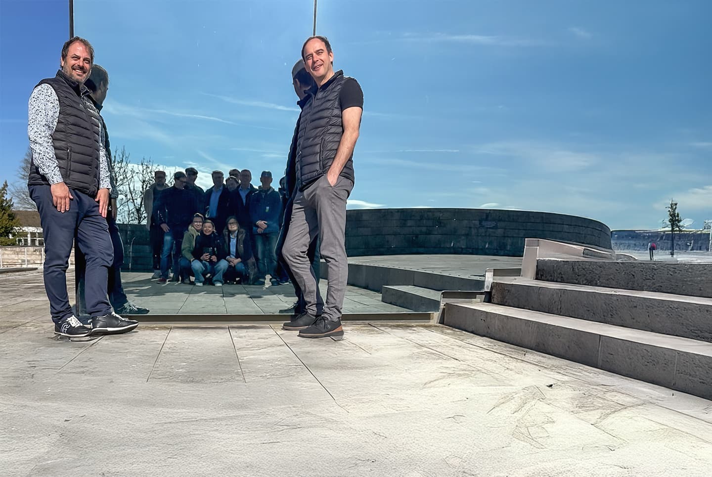 Das CombiTeam sitzt auf dem Dach der Grimmwelt in Kassel vor einem Spiegel, sodass dieses nur im Spiegel zu sehen ist. Vor dem Spiegel stehen die Geschäftsführer die CombiBrüder Stefan und Sebastian Grimm, beide lächeln in die Kamera. Der Himmel ist blau und das Wetter scheint schön zu sein. 