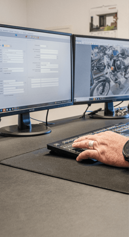 Das Bild zeigt einen Kfz-Sachverständigen, der mit der Software CombiPlus Professional auf zwei Monitoren arbeitet. Auf dem linken Bildschirm ist eine Benutzeroberfläche der Software zu sehen, die verschiedene Felder zur Eingabe von Fahrzeug- und Schadendaten enthält. Der rechte Bildschirm zeigt ein Bild von Motorrädern, das wahrscheinlich zur Schadenbewertung dient. Die Hand des Sachverständigen, die auf der Tastatur liegt, verdeutlicht die Interaktivität mit der Software für die Erstellung der Gutachten. Diese Darstellung unterstreicht, wie CombiPlus Professional den Sachverständigen bei der präzisen und schnellen Erstellung von Kfz-Gutachten unterstützt.