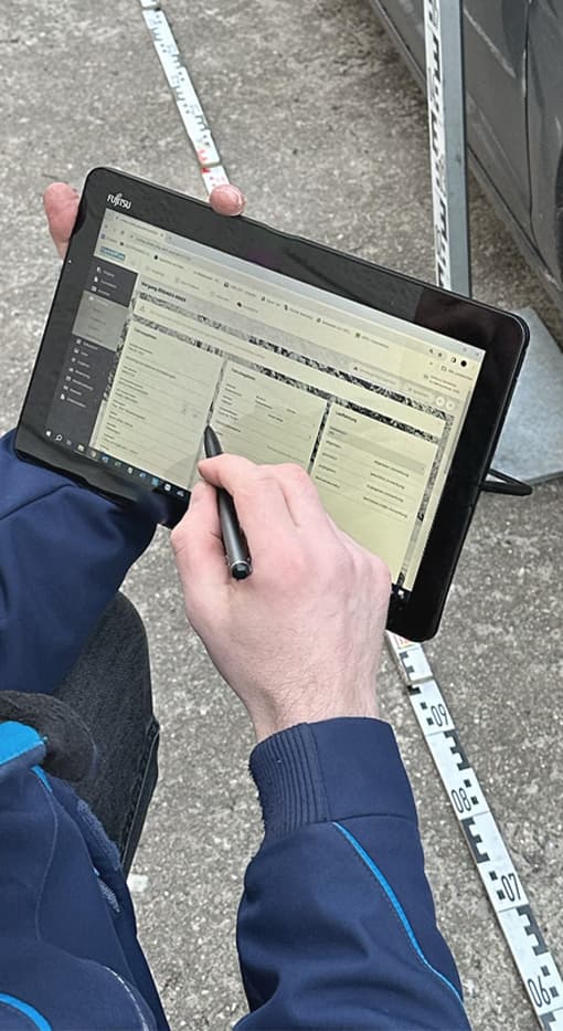 Das Bild zeigt einen Kfz-Sachverständigen, der mit einem Tablet arbeitet, auf dem die CombiPlus Online Software für die mobile Schadenaufnahme verwendet wird. Der Sachverständige hält das Tablet mit einer Hand und nutzt einen Stift, um auf der Bildschirmoberfläche zu navigieren. Im Hintergrund ist ein Maßband sichtbar, da der Sachverständige direkt vor Ort am Fahrzeug arbeitet und die Schäden präzise dokumentiert. Diese mobile Lösung ermöglicht eine schnelle und effiziente Schadenaufnahme und -bearbeitung direkt beim Kunden vor Ort.