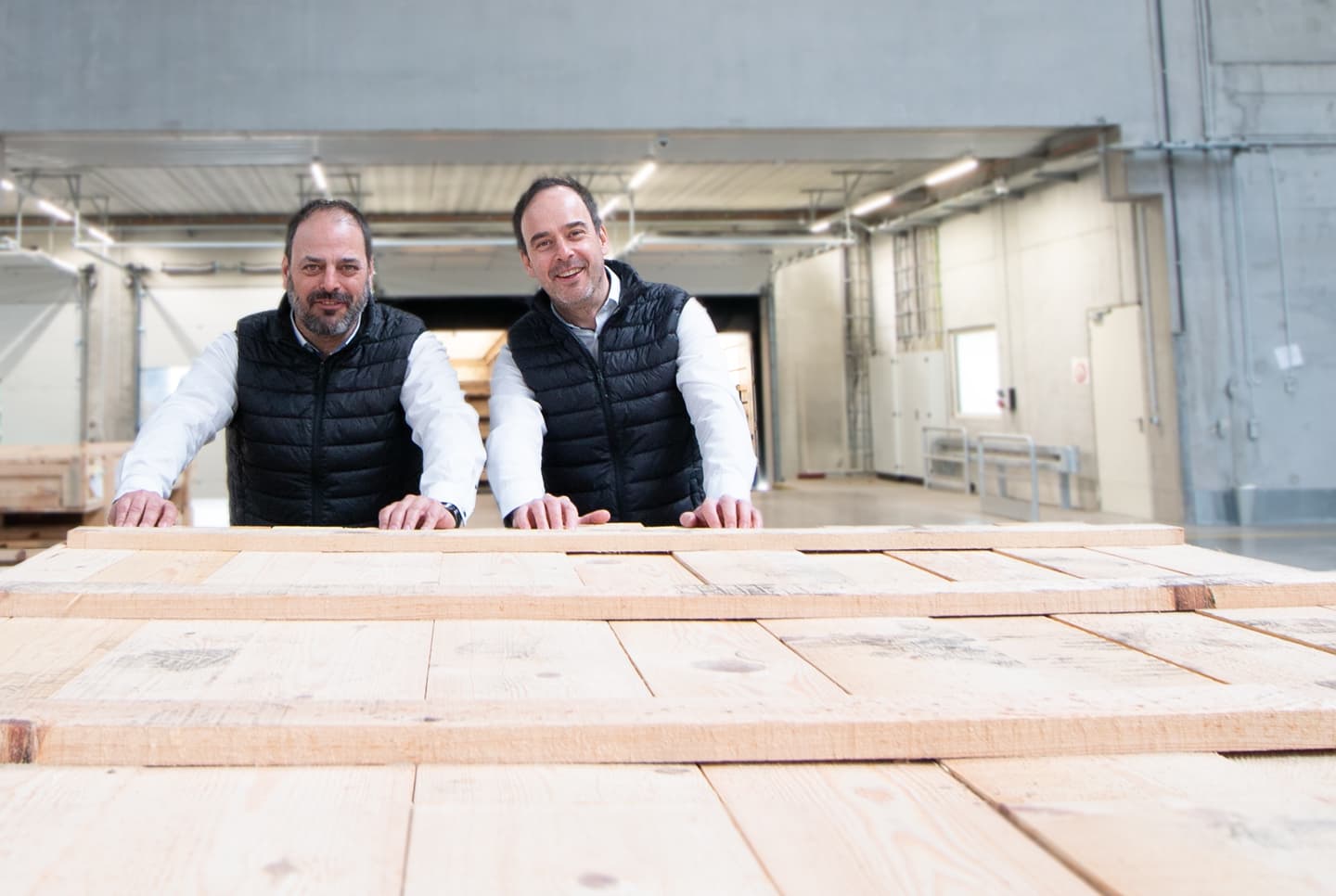 Die CombiBrüder Grimm, Sebastian und Stefan Grimm, Geschäftsführer von CombiSystems, stehen in einer großen, modernen Lagerhalle hinter einer Holzpalette. Beide tragen weiße Hemden und schwarze Steppwesten. Ihr freundliches und engagiertes Auftreten unterstreicht ihre Rolle als Führungspersönlichkeiten. Im Hintergrund ist die offene Halle mit industriellem Ambiente und natürlichem Licht zu sehen, was die Verbindung von Professionalität und Praxisnähe vermittelt.