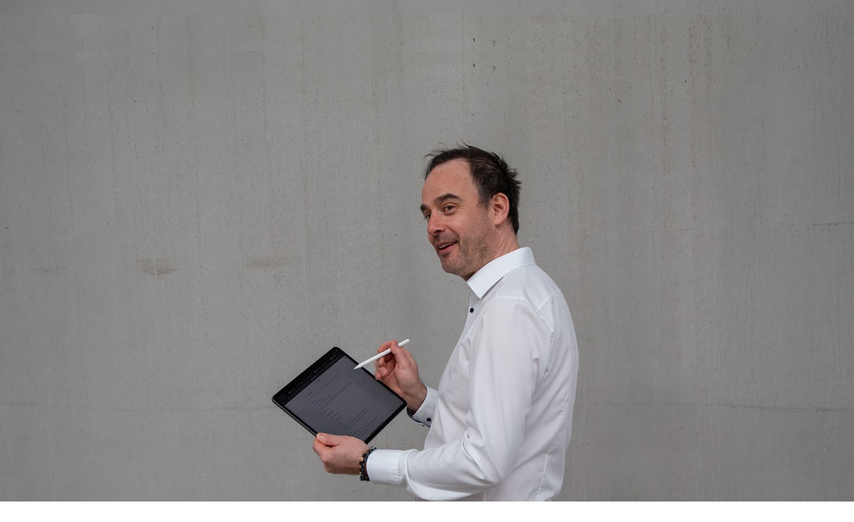 Sebastian Grimm, Geschäftsführer bei CombiSystems, steht vor einer schlichten grauen Wand. Er trägt ein weißes Hemd und hält ein Tablet in der Hand, während er mit einem Stift darauf schreibt. Sein Blick ist freundlich, und er wirkt konzentriert und engagiert. Die minimalistische Umgebung lenkt den Fokus auf seine professionelle Ausstrahlung und den Einsatz moderner Technologien.
