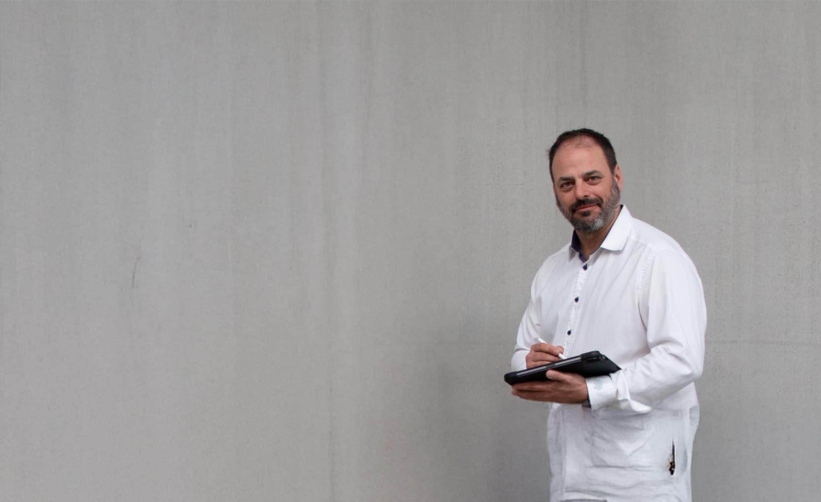 Stefan Grimm, Geschäftsführer bei CombiSystems, steht vor einer schlichten grauen Wand. Er trägt ein weißes Hemd und hält ein Tablet in der Hand, während er lächelnd in die Kamera blickt. Sein Auftreten wirkt professionell und freundlich. Die minimalistische Umgebung lenkt den Fokus auf seine Person und vermittelt einen modernen, geschäftlichen Kontext.