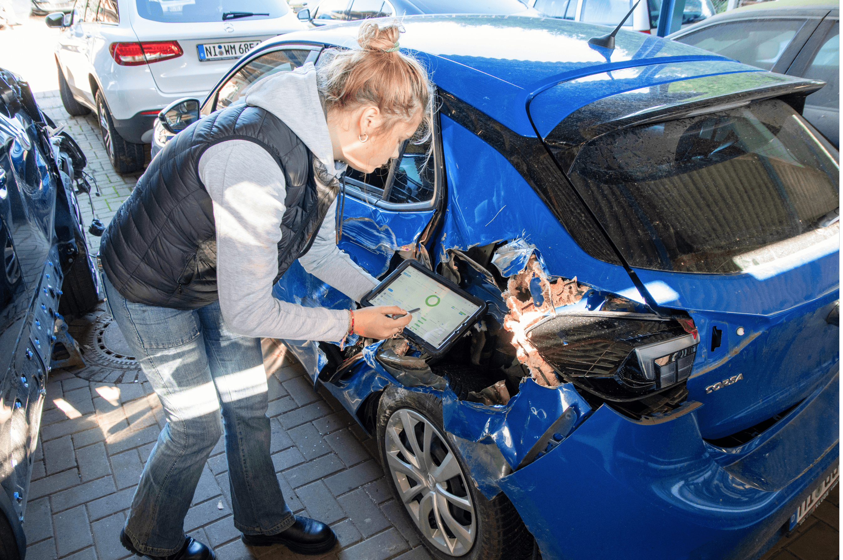 Das Bild zeigt eine Frau, die ein Tablet in den Händen hält, während sie den Schaden an einem blauen Auto inspiziert. Der Schaden an der linken Fahrzeugseite ist deutlich sichtbar, insbesondere an der hinteren Tür und dem Rücklicht. Auf dem Tablet-Bildschirm ist eine Schadenbewertung sichtbar, die auf eine Erfassung des Schadens in einem System hinweist. Im Hintergrund sind weitere Fahrzeuge zu sehen, die den Kontext einer Werkstattumgebung suggerieren.