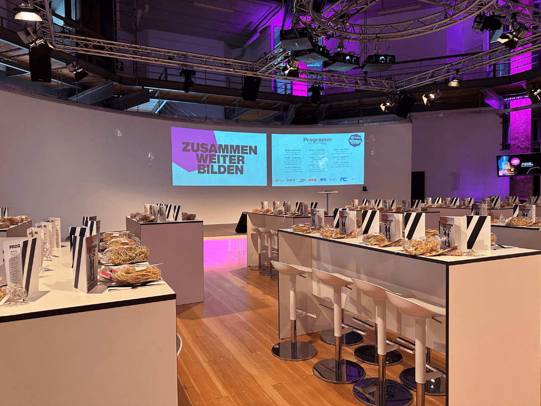 Der Veranstaltungsraum des CombiBildungsBuffets im Oktogon Essen ist stilvoll vorbereitet. Hohe Tische mit weißen Barhockern sind mit Infomaterialien, Geschenktüten und kulinarischen Snacks gedeckt. Im Hintergrund zeigt eine große Leinwand das Motto 'Zusammen Weiter Bilden' sowie das Veranstaltungsprogramm. Die Bühne ist beleuchtet, während das Ambiente durch violette Akzentlichter hervorgehoben wird