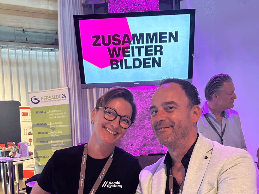 Acoll Busche und Sebastian Grimm lächeln in die Kamera beim CombiBildungsBuffet in Essen. Im Hintergrund ist ein Bildschirm mit dem Schriftzug 'Zusammen Weiter Bilden' sowie verschiedene Messestände zu sehen.