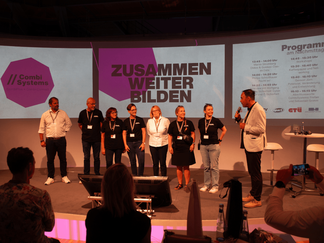 Das CombiTeam steht auf der Bühne des CombiBildungsBuffets vor einer großen Leinwand mit dem Schriftzug 'Zusammen Weiter Bilden'. Sebastian Grimm spricht zum Team, während das Veranstaltungsprogramm auf der rechten Seite der Leinwand angezeigt wird.