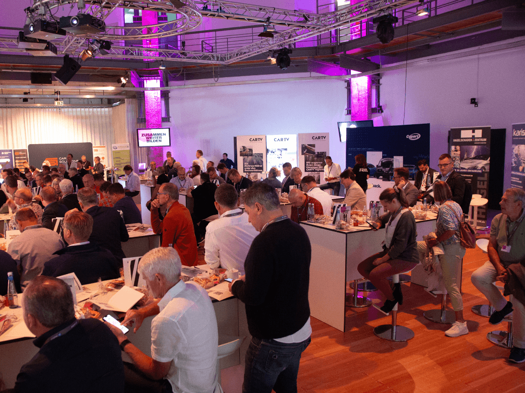 Teilnehmer des CombiBildungsBuffets sitzen und stehen an Tischen in einem großen Veranstaltungsraum mit moderner Beleuchtung. Die Atmosphäre ist lebendig, während die Gäste sich austauschen, essen und Notizen machen. Im Hintergrund sind Messe- und Informationsstände verschiedener Unternehmen zu sehen, sowie eine Leinwand mit der Aufschrift 'ZUSAMMEN WEITER BILDEN'.