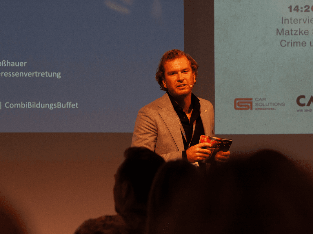 Philipp Schloßhauer steht auf einer Bühne und hält einen Vortrag beim CombiBildungsBuffet. Er trägt ein graues Sakko über einem schwarzen Hemd und spricht mit einem Headset-Mikrofon. In seiner Hand hält er ein rotes Notizbuch oder eine Broschüre. Der Hintergrund zeigt eine große Leinwand mit Logos und Veranstaltungstext.