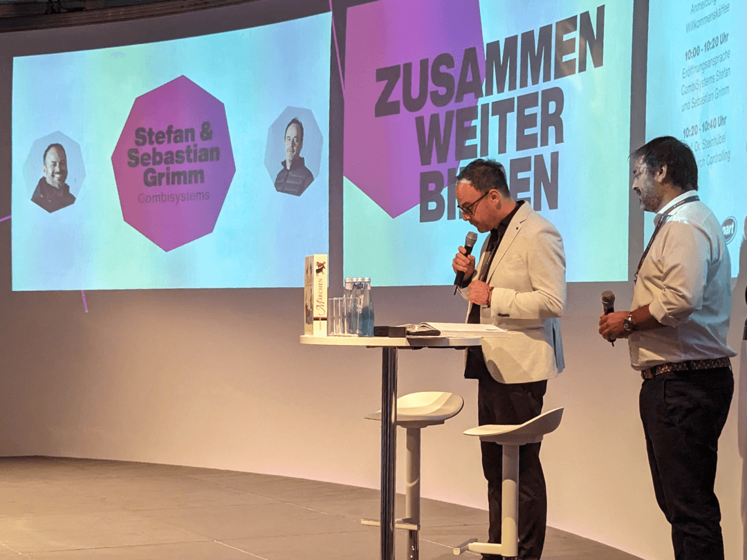 Bühne des CombiBildungsBuffets mit einer großen Leinwand im Hintergrund, auf der die Worte 'ZUSAMMEN WEITER BILDEN' sowie die Namen 'Stefan & Sebastian Grimm' mit ihren Porträts zu sehen sind. Zwei Moderatoren stehen an einem hohen Tisch mit Mikrofonen in der Hand, während auf dem Tisch Wasserflaschen und eine Geschenkbox stehen. 