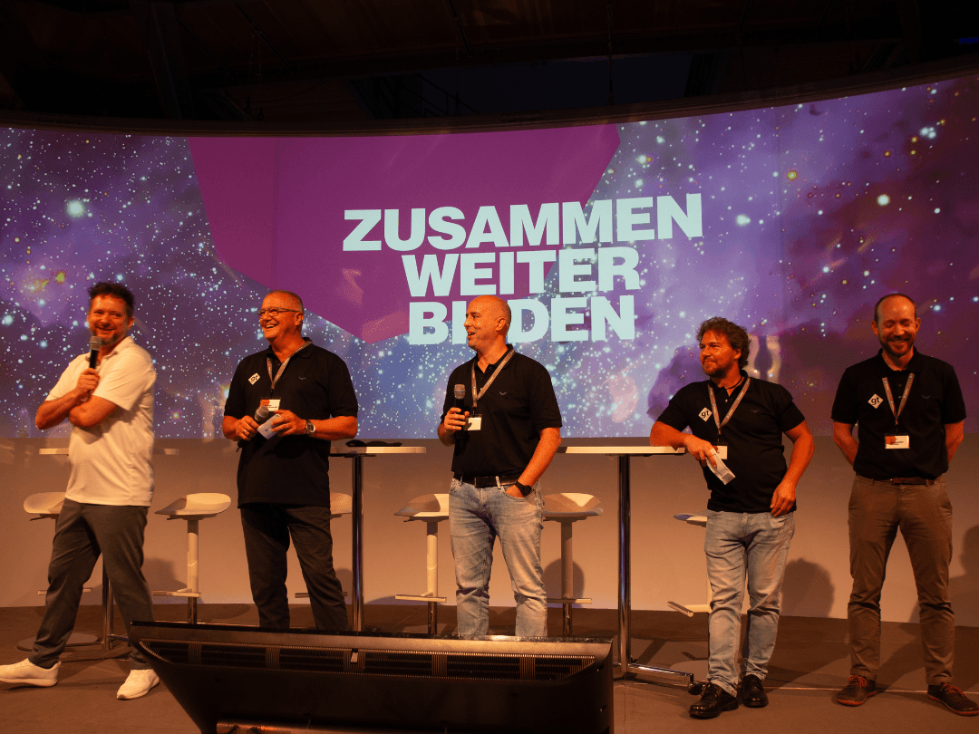 Szene vom letzten CombiBildungsBuffet: Fünf Männer stehen auf einer Bühne vor einer großen Leinwand mit dem Schriftzug 'ZUSAMMEN WEITER BILDEN'. Der Hintergrund zeigt eine galaxieartige Darstellung mit violetten und blauen Farbtönen. Einer der Männer hält ein Mikrofon und spricht, während die anderen zuhören und lächeln. Sie tragen schwarze Poloshirts mit einem Firmenlogo, bis auf einen, der in einem weißen Shirt gekleidet ist. Im Vordergrund ist ein Monitor zu sehen.