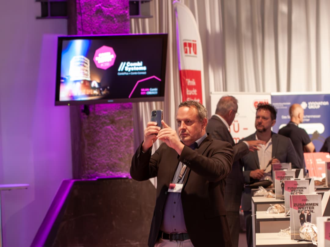  ChatGPT: Auf dem Bild ist Bernd Antoni zu sehen, wie er mit seinem Smartphone eine Aufnahme macht. Im Hintergrund befinden sich verschiedene Messestände, Banner von GTÜ und Innovation Group sowie ein Bildschirm mit dem CombiBildungsBuffet-Design. Zudem sind mehrere Teilnehmer im Gespräch, darunter auch Gereon Jörn, der als Speaker aufgetreten ist.