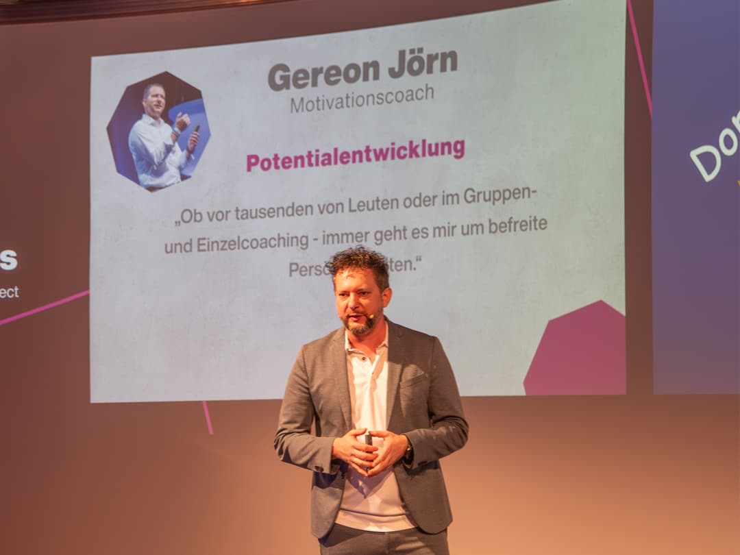 Das Bild zeigt den Motivationscoach Gereon Jörn während seines Vortrags zum Thema Potenzialentwicklung auf dem CombiBildungsBuffet. Er spricht vor einer großen Leinwand, auf der sein Name, Titel und ein Zitat zu seiner Arbeit zu sehen sind. Der Vortrag befasst sich mit persönlicher Weiterentwicklung und Coaching.