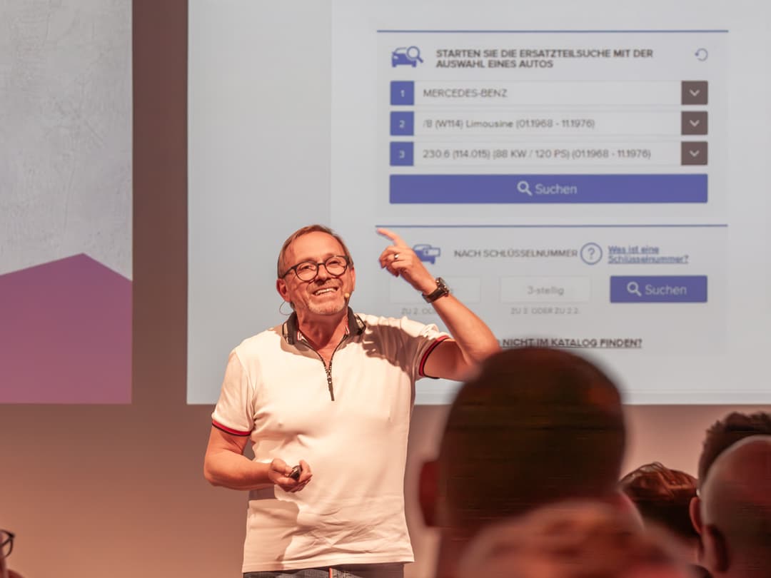 Martin Stromberg steht auf der Bühne und hält einen Vortrag. Er trägt ein weißes Polo-Shirt mit roten Akzenten und eine Brille. Mit einem Lächeln deutet er auf die große Präsentationsfolie hinter ihm, auf der eine digitale Benutzeroberfläche zur Ersatzteilsuche für Autos zu sehen ist. Das Publikum verfolgt aufmerksam die Präsentation
