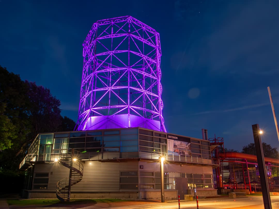 Das Oktogon in Essen bei Nacht: Ein modern umgebautes Industriebauwerk mit einem markanten stählernen Turm, der in leuchtendem Lila angestrahlt wird. Die untere Gebäudestruktur besteht aus Glas- und Metallelementen, während eine Wendeltreppe an der Seite nach oben führt. Der dunkle Nachthimmel bildet einen starken Kontrast zur futuristischen Beleuchtung.