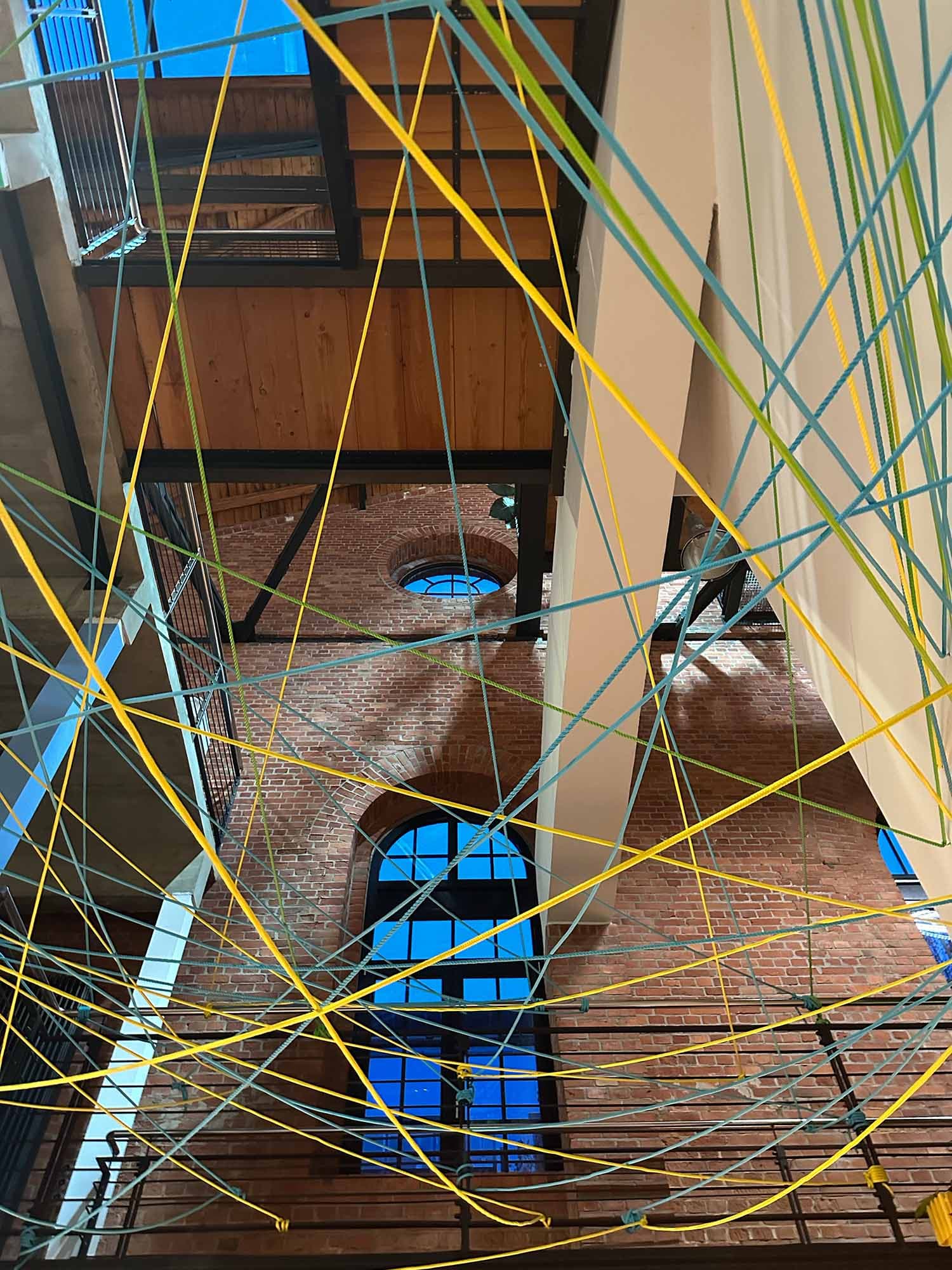 Blick nach oben in einem offenen Backsteingebäude mit hohen Fenstern, durchzogen von gelben und blauen gespannten Seilen. Das Netz symbolisiert Verbindungen und Vernetzung, passend zur Atmosphäre des MachWerks.