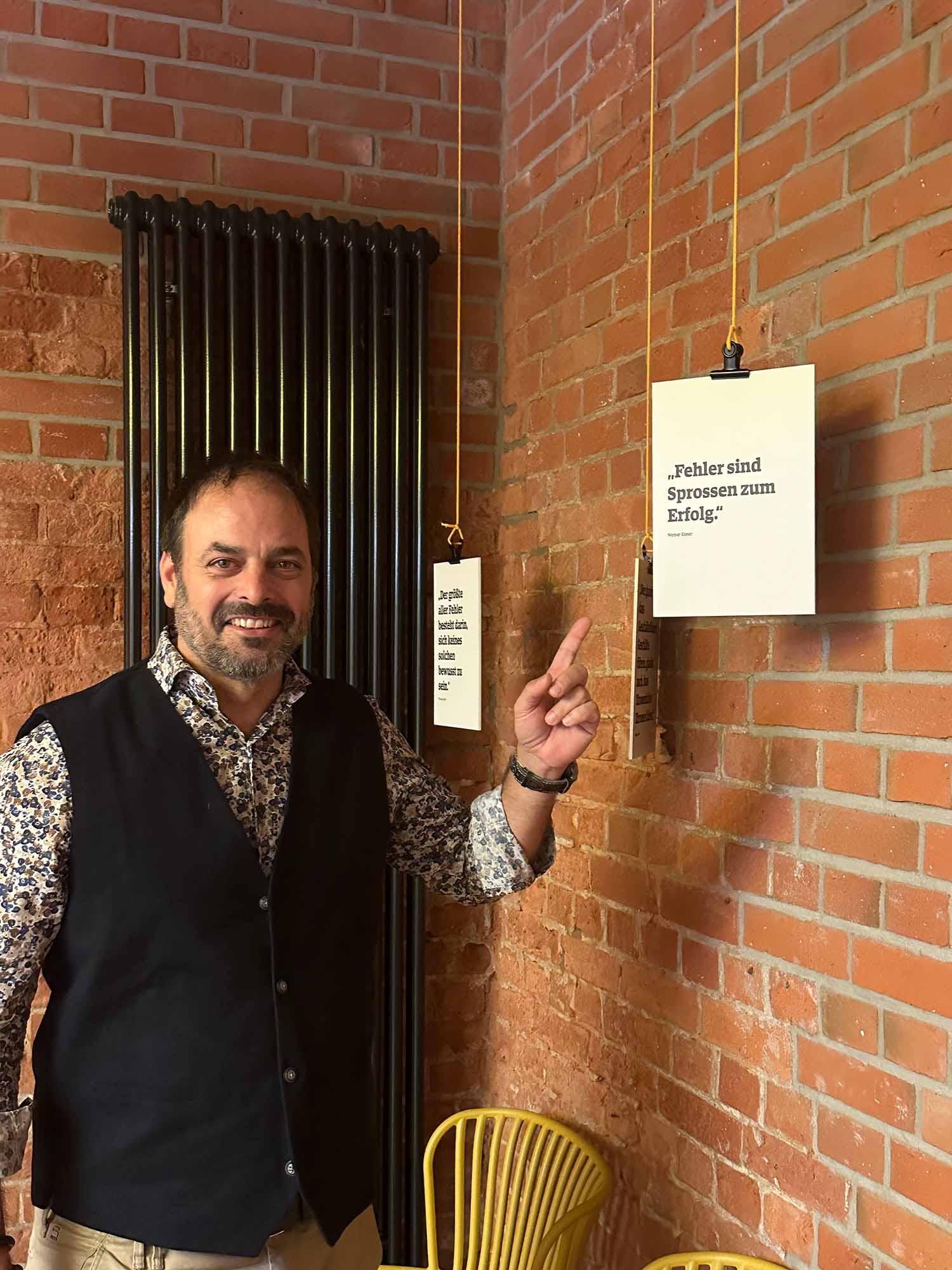 Stefan Grimm steht in einem Raum mit Backsteinwänden im MachWerk und zeigt lächelnd auf ein Zitat an der Wand: 'Fehler sind Sprossen zum Erfolg.' Im Hintergrund hängen weitere motivierende Zitate an gelben Schnüren, ein schwarzer Heizkörper und gelbe Stühle ergänzen das Ambiente.