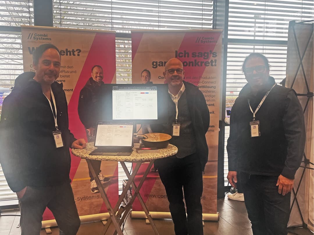 Das CombiTeam beim HagelCampus: Sebastian Grimm, Christian Schmidt und Stefan Grimm stehen an einem Messestand mit zwei Laptops und Roll-Ups im Hintergrund, die die CombiPlus- und CombiConnect-Lösungen präsentieren. Alle drei tragen Namensschilder und lächeln in die Kamera. Der Stand befindet sich vor großen Fensterfronten mit Lamellen.