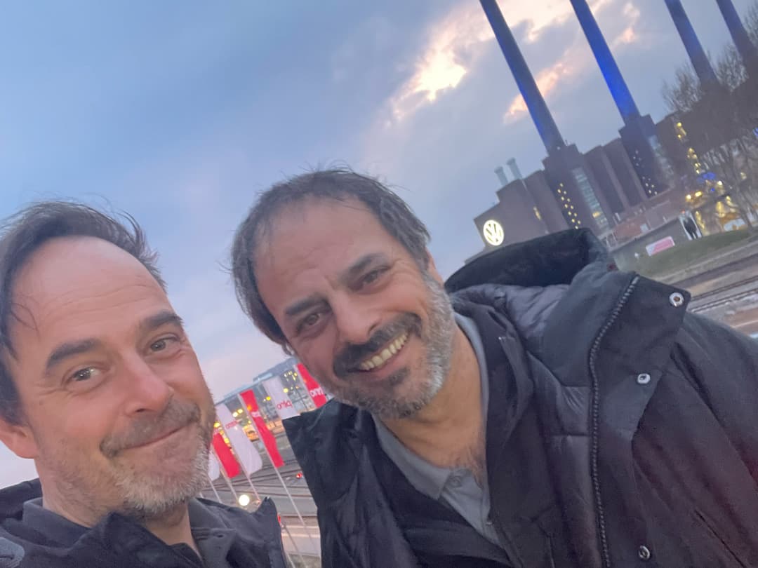 Sebastian und Stefan Grimm machen ein Selfie beim HagelCampus in Wolfsburg. Im Hintergrund ist das VW-Werk mit seinen markanten Schornsteinen und dem leuchtenden Volkswagen-Logo zu sehen. Der Himmel ist leicht bewölkt, und beide tragen dunkle Jacken und lächeln in die Kamera.
