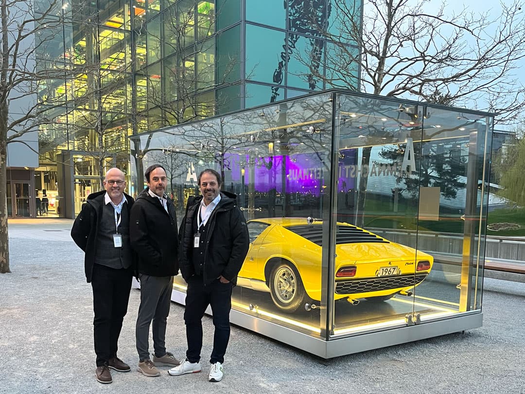 Stefan Grimm, Sebastian Grimm und Christian Schmidt vom CombiTeam stehen vor einem gläsernen Schaukasten mit einem gelben Lamborghini Miura von 1967 in der Autostadt Wolfsburg. Hinter ihnen leuchtet ein modernes Gebäude mit bunter Beleuchtung. Alle drei tragen Namensschilder und lächeln in die Kamera.