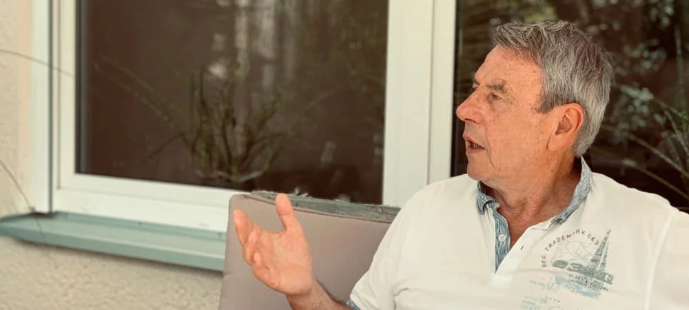 Bodo Grimm im Sommerinterview, gestikulierend und im Gespräch vertieft – aufgenommen auf der Terrasse mit Blick auf das Fenster im Hintergrund.