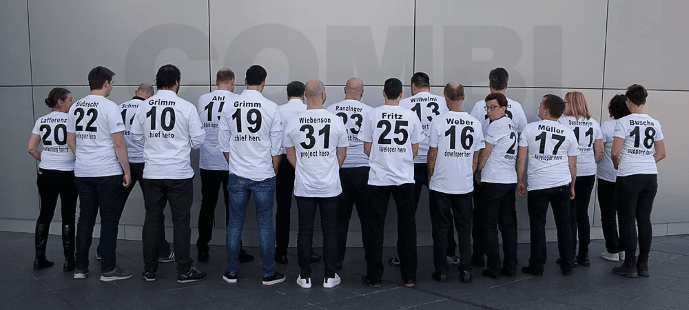 das CombiTeam in einer wunderbar inszenierten Gruppenaufnahme von hinten, alle mit individuellen Team-T-Shirts. Auf den Rücken der weißen Shirts stehen jeweils ein Name, eine Nummer und eine Rolle im Team wie z. B. „developer hero“, „chief here“, „project hero“ oder „support hero“.