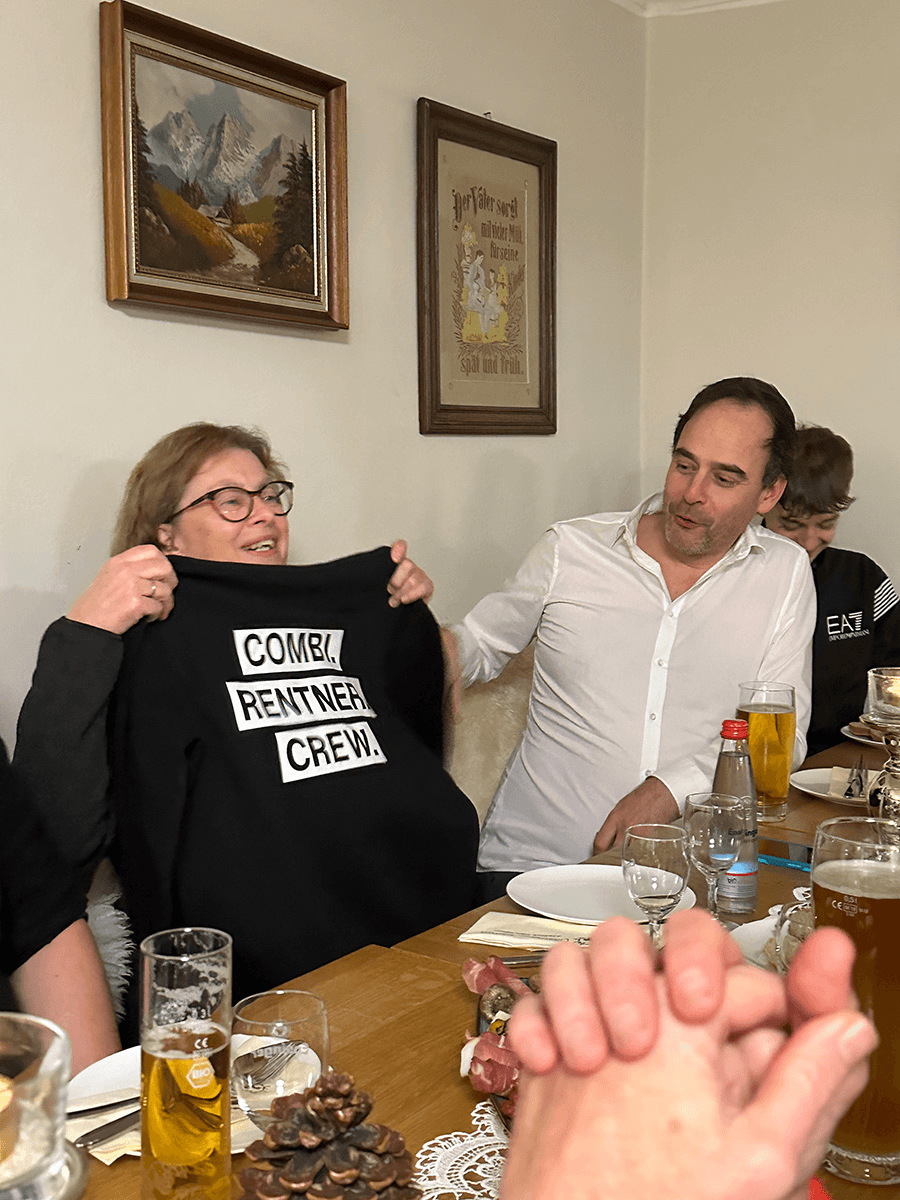  Auf dem Bild sieht man Käthe Pegel und Sebastian Grimm bei einer feierlichen Zusammenkunft. Käthe hält stolz ein schwarzes Sweatshirt mit der Aufschrift „COMBI RENTNER CREW.“ in die Höhe – offenbar ein Geschenk zur Verabschiedung in den Ruhestand. Sebastian sitzt lächelnd neben ihr, was auf eine herzliche und wertschätzende Atmosphäre im Team hindeutet.