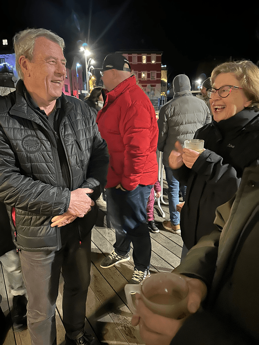 Käthe Pegel und Bodo Grimm bei einem geselligen Abend unter freiem Himmel – vermutlich bei einem winterlichen Event oder Weihnachtsmarkt. Beide wirken gut gelaunt im Gespräch, mit Heißgetränken in der Hand und warm eingepackt.