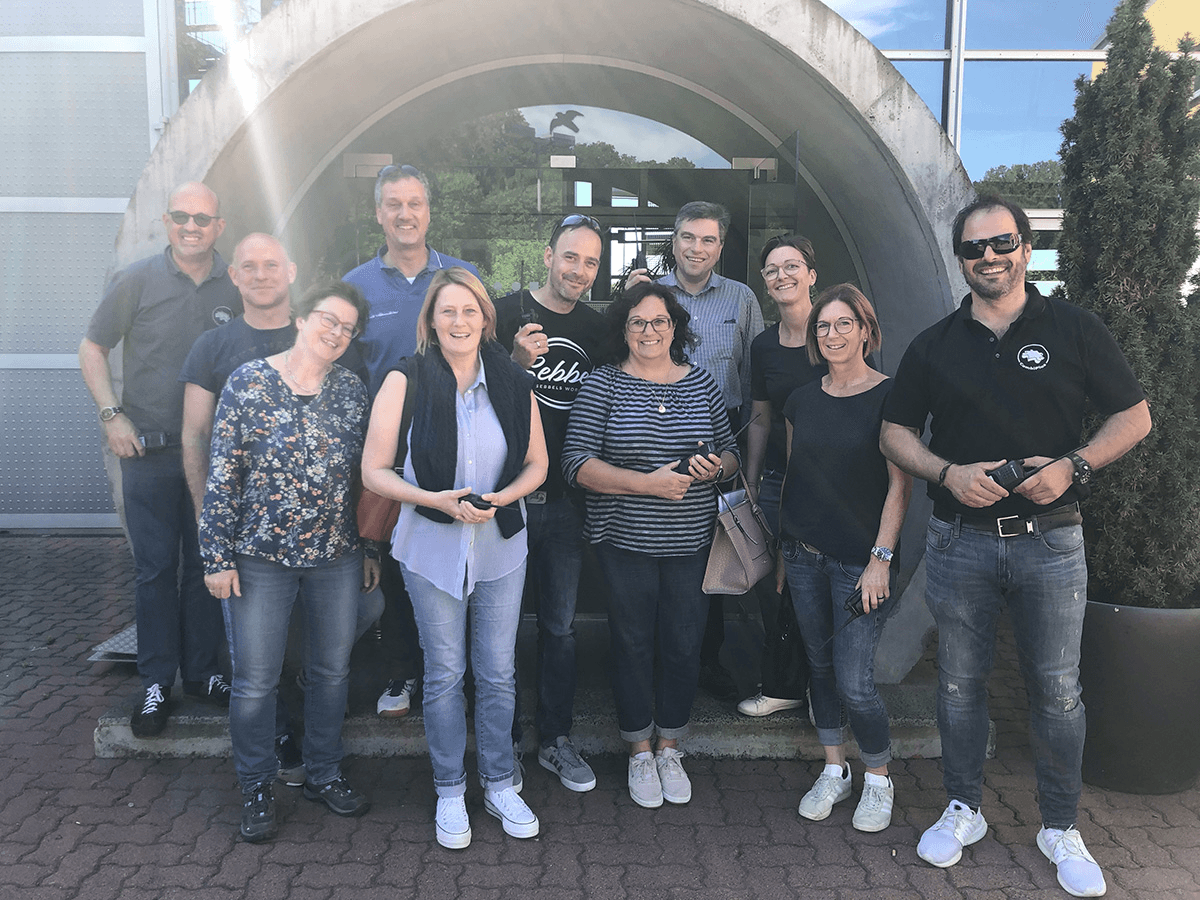 Gruppenfoto des CombiTeams vor einem modernen Gebäude mit Rundbogen-Eingang – lächelnde Kolleginnen und Kollegen bei sonnigem Wetter und entspannter Stimmung.