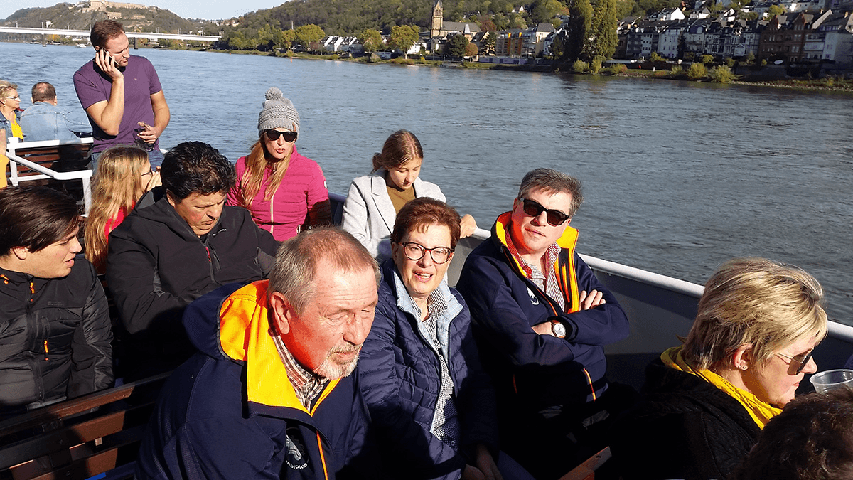 Das CombiTeam auf einem Ausflugsschiff bei sonnigem Wetter – entspannt sitzend an Deck mit Blick auf den Rhein und eine malerische Altstadt im Hintergrund.