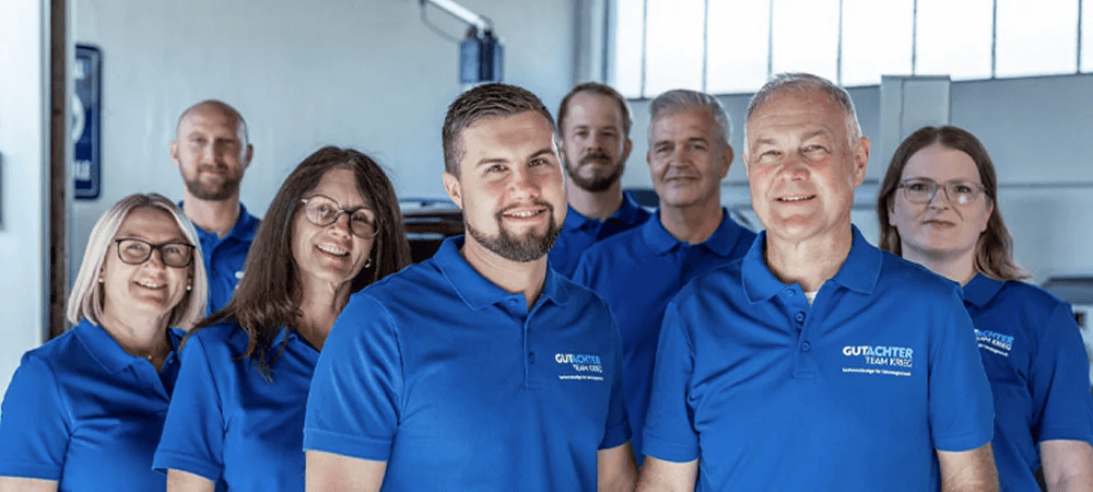 Gruppenfoto des Gutachter Team Krieg: Acht Teammitglieder stehen in einer Werkstatt, tragen einheitliche blaue Poloshirts mit Firmenlogo und lächeln in die Kamera – ein sympathisches und professionelles Team, das für Kompetenz im Kfz-Schadenmanagement steht.