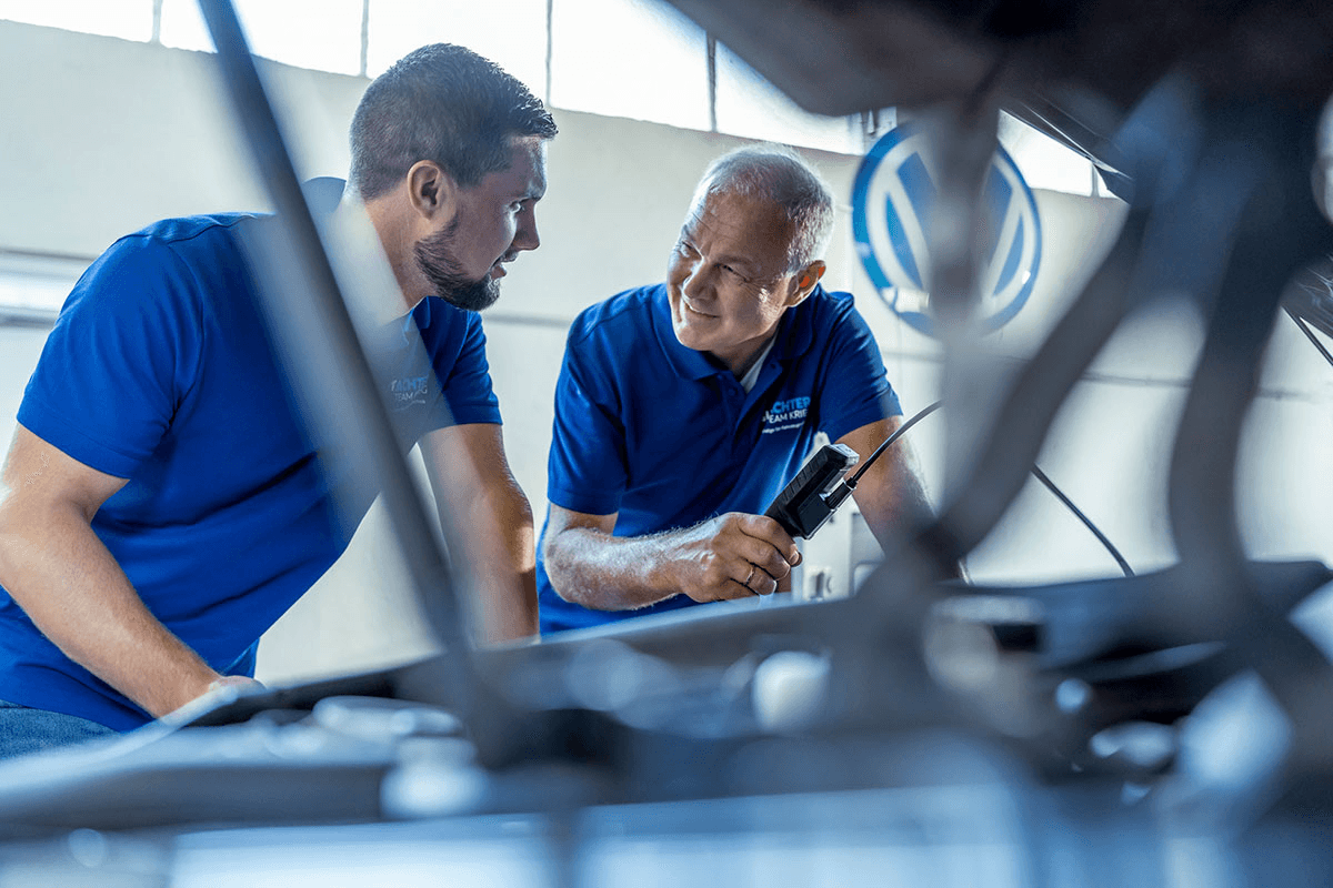 Heiko und Piere Krieg vom Gutachter Team Krieg in blauen Poloshirts beugen sich gemeinsam über den geöffneten Motorraum eines Fahrzeugs. Einer von ihnen hält ein Diagnosegerät in der Hand, beide wirken konzentriert im Gespräch. Die Szene vermittelt Teamarbeit und technisches Know-how.
