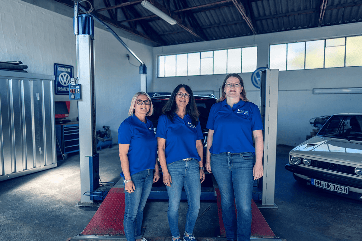 Drei Frauen vom Gutachter Team Krieg stehen nebeneinander in einer Werkstatt, alle in blauen Team-Shirts mit Logo. Im Hintergrund sind eine Hebebühne, Werkstatteinrichtung und ein klassischer VW zu sehen. Die Atmosphäre wirkt professionell und freundlich.