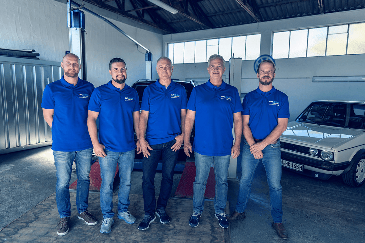 Das Team von Gutachter Krieg: Fünf Männer in einheitlichen blauen Polo-Shirts mit "Gutachter Krieg" Logo stehen nebeneinander in einer Werkstatt. Im Hintergrund befinden sich ein schwarzes Fahrzeug auf einer Hebebühne und ein weißer Oldtimer (VW Golf). Die Werkstatt ist lichtdurchflutet mit hoher Decke, sichtbaren Stahlträgern und großen Fenstern.