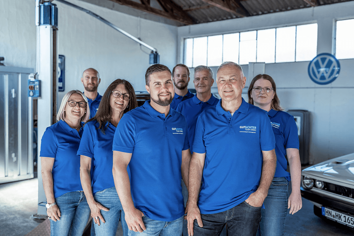 Das erweiterte Team von Gutachter Krieg: Acht Teammitglieder – fünf Männer und drei Frauen – tragen einheitlich blaue Polo-Shirts mit dem Logo „Gutachter Team Krieg“. Im Vordergrund stehen Pierre Krieg und Heike Krieg. Die Gruppe posiert in einer Werkstatt mit sichtbaren Hebebühnen, großem VW-Logo an der Wand und einem klassischen VW Golf. Die Atmosphäre ist professionell und freundlich.