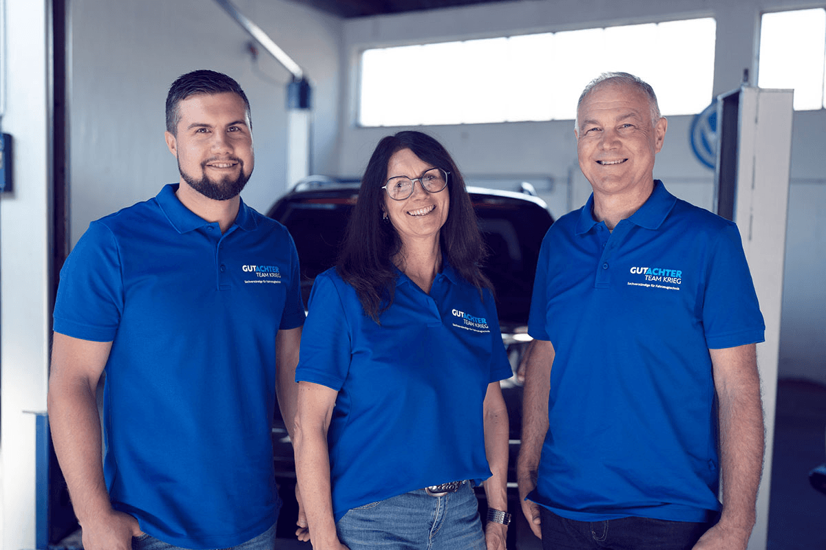 Pierre Krieg, Heiko Krieg und ein weiterer Kollege vom Gutachter Team Krieg stehen nebeneinander in einer Werkstatt. Alle drei tragen einheitliche blaue Polo-Shirts mit dem Logo „Gutachter Team Krieg“. Im Hintergrund ist ein schwarzes Fahrzeug und ein Werkstattfenster mit diffusem Tageslicht zu sehen. Alle lächeln und wirken freundlich und professionell.