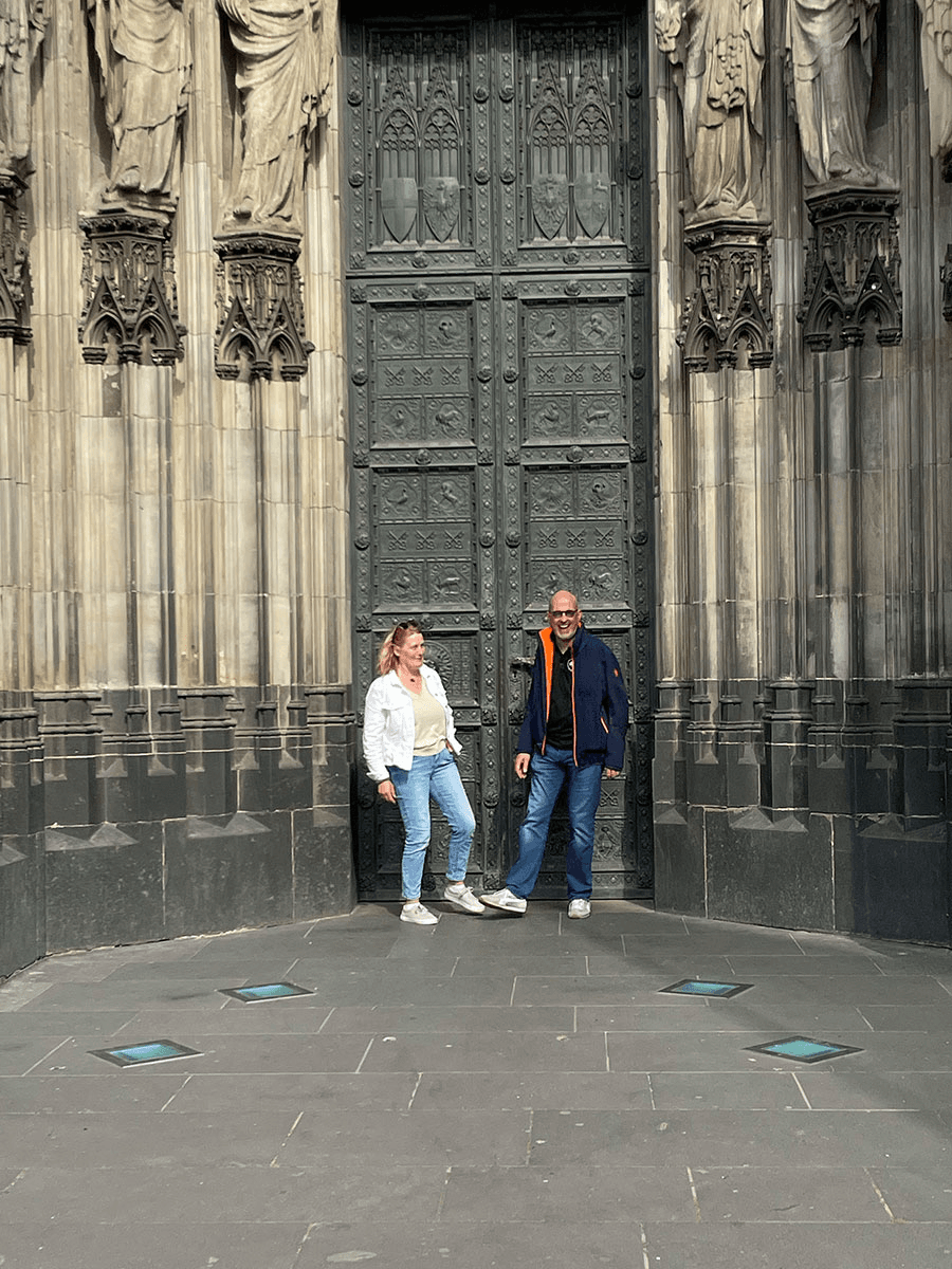 Maren Sundermeyer und Christian Schmidt vom CombiTeam stehen vor dem imposanten Hauptportal des Kölner Doms. Die beiden genießen einen sonnigen Moment vor der detailreich verzierten gotischen Tür.