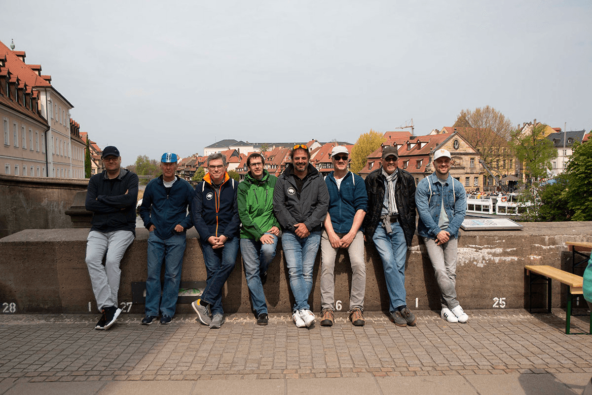 Das CombiTeam sitzt entspannt auf einer Steinmauer vor der malerischen Kulisse mit Fachwerkhäusern, roten Dächern und frühlingshaftem Grün – ein Moment des Teamgeists und der Verbundenheit in historischer Umgebung.