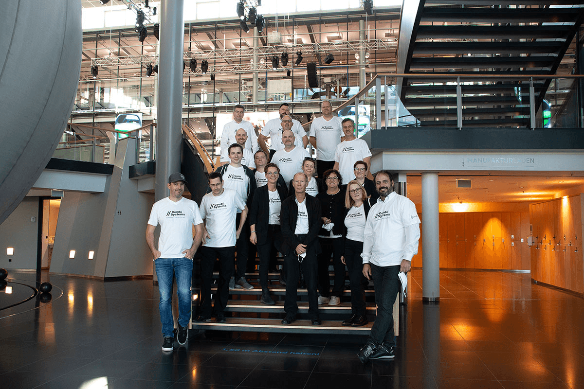 Das CombiTeam versammelt sich auf einer Treppe im modernen Foyer der Autostadt Wolfsburg – alle tragen weiße CombiSystems-Shirts und strahlen Teamgeist, Zusammenhalt und Begeisterung für gemeinsame Ziele aus.
