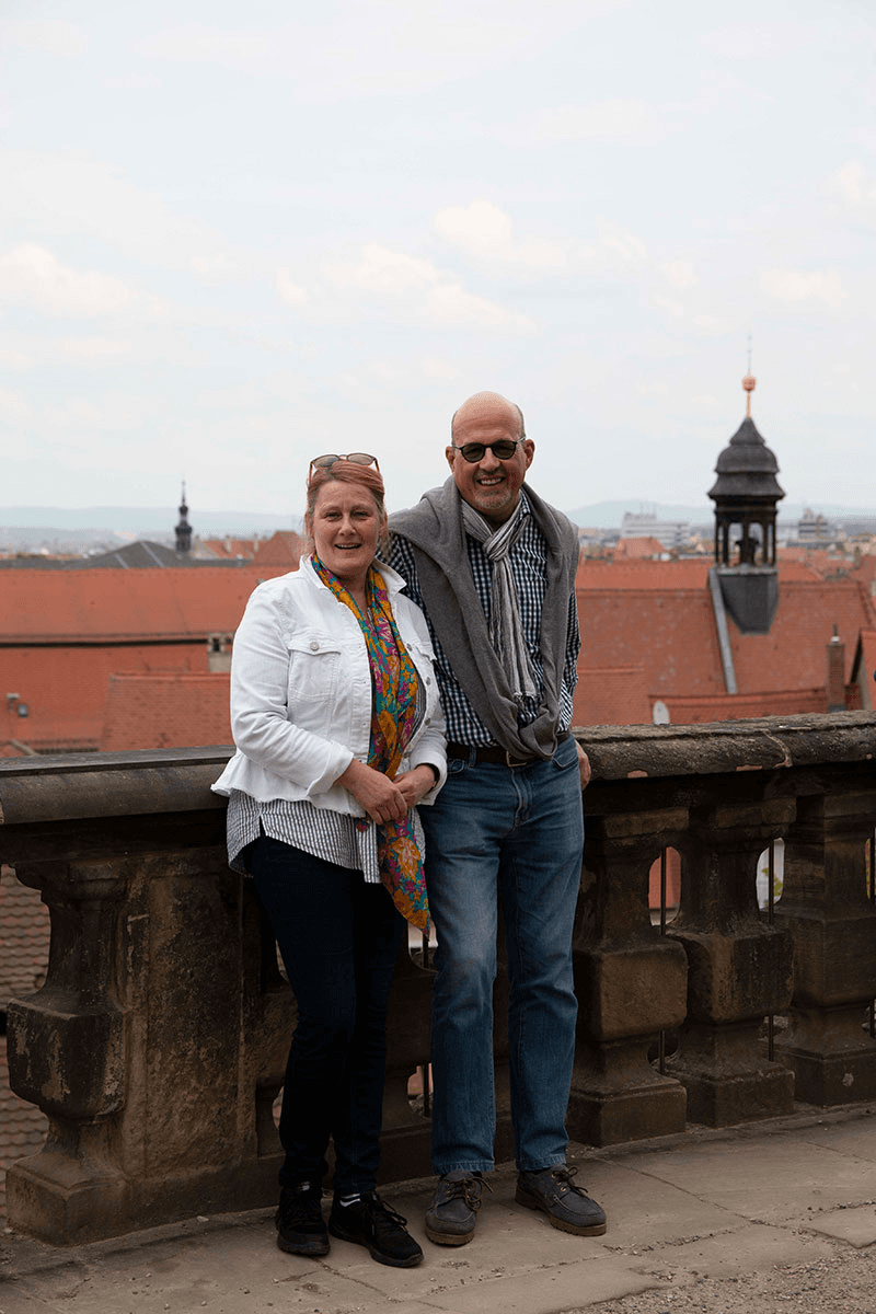 Maren Sundermeyer und Christian Schmidt stehen lächelnd vor einer historischen Brüstung mit Blick über die roten Dächer einer Altstadt – ein Moment der Ruhe und des Zusammenhalts im CombiTeam.