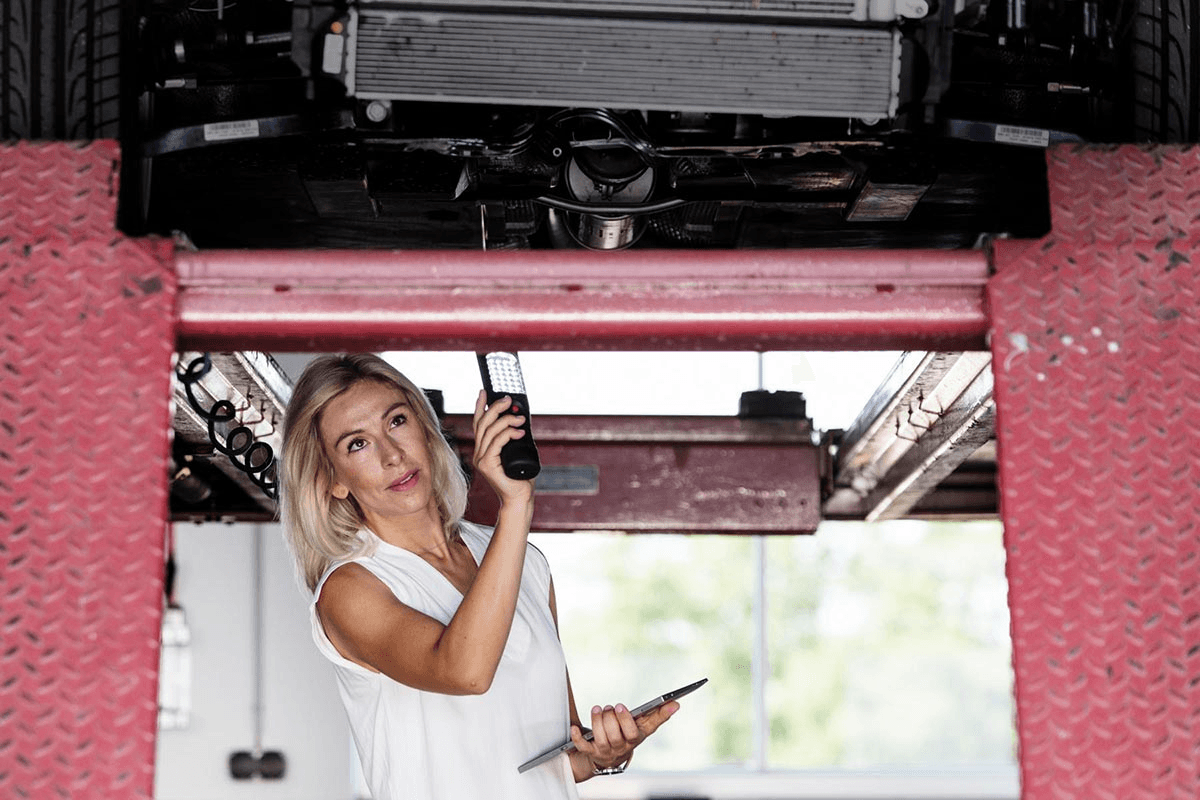 Victoria von Alkewitz bei der Begutachtung eines Fahrzeugs in einer Werkstatt. Sie inspiziert aufmerksam den Unterboden eines Autos auf einer Hebebühne mit einer Arbeitslampe in der Hand 