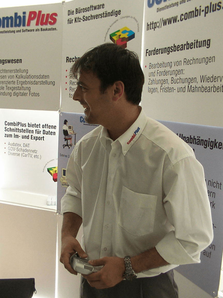 Auf dem Bild trägt Stefan Grimm ein weißes, langärmeliges Hemd mit dem combi.plus-Logo am Kragen. Die Ärmel sind hochgekrempelt. Er hält eine silberne Digitalkamera in den Händen und trägt eine silberne Armbanduhr am linken Handgelenk. Dazu kombiniert er eine graue Stoffhose. Das Gesamtbild wirkt gepflegt, professionell und gleichzeitig bodenständig – passend zu einem Messeauftritt.