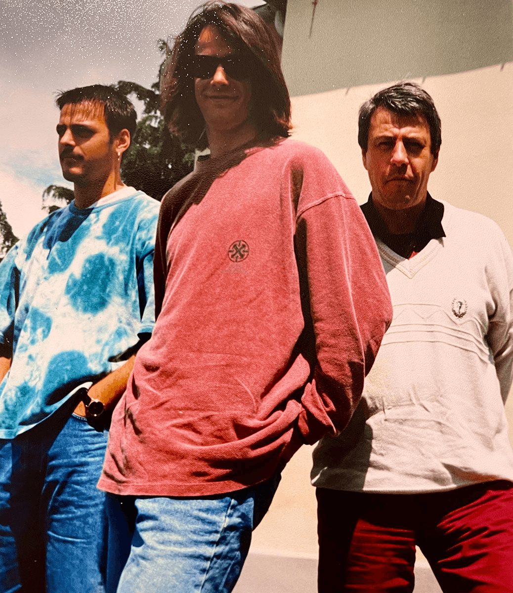 Ein nostalgisches Foto aus den frühen Jahren zeigt Bodo Grimm, Stefan Grimm und Sebastian Grimm entspannt im Freien. Sebastian steht mittig mit langen Haaren und Sonnenbrille, lässig gekleidet in einem weinroten Pullover mit Logo. Stefan trägt ein türkis-blaues Batik-Shirt und eine Armbanduhr, während Bodo rechts im Bild in einem beigen Pullover mit V-Ausschnitt zu sehen ist. Die drei wirken gelöst und verkörpern einen typischen Stil der 90er Jahre – ein authentischer Rückblick auf die Anfänge des späteren CombiSystems