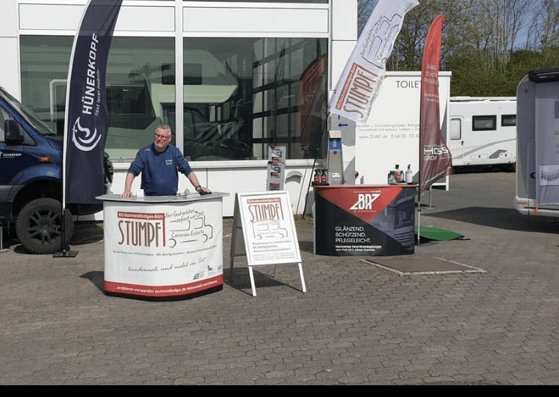 Rainer Stumpf am Messestand seines Kfz-Sachverständigenbüros im Außenbereich einer Caravan- oder Fahrzeugmesse. Neben dem Infostand mit dem Logo „STUMPF Caravan-Experten“ befinden sich Flaggen, Infotafeln und weitere Aussteller, darunter ein Stand für Fahrzeugpflegeprodukte. Im Hintergrund sind Wohnmobile und ein moderner Ausstellungsbereich zu sehen – eine Szene, die seine mobile Gutachtertätigkeit und Branchennähe unterstreicht.