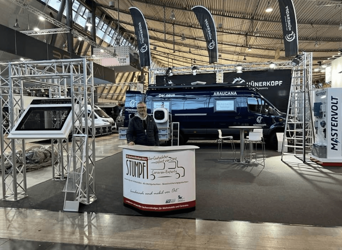 Rainer Stumpf am Stand seines Kfz-Sachverständigenbüros „Stumpf Caravan-Experten“ auf einer Fachmesse für Caravaning und Fahrzeugtechnik. Der Messestand ist modern gestaltet mit einem auffälligen Infodesk im Vordergrund, einem stylischen blauen Van im Hintergrund und verschiedenen technischen Präsentationen rund um Wohnmobile. Die Szene zeigt seine Spezialisierung auf mobile Gutachten und Beratung rund um Reisemobile.