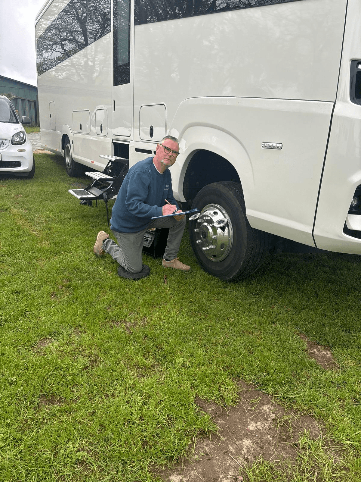 Rainer Stumpf kniet auf einer Wiese vor einem hochwertigen Wohnmobil und dokumentiert den Zustand des Fahrzeugs mit einem Klemmbrett. Er trägt eine grüne Arbeitshose, beigen Schuhe und ein dunkelblaues Sweatshirt mit der Aufschrift „SV Büro Stumpf“. Im Hintergrund ist ein Smart zu sehen. Die Szene zeigt den mobilen Einsatz als Kfz-Sachverständiger bei der Begutachtung vor Ort.